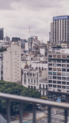 A cityscape featuring mid-rise buildings, with a mix of modern and older architecture. The scene includes a prominent bank building displaying signage and various residential or office blocks, some showing wear. Below, a busy street intersection is partially visible, bordered by lush green trees.