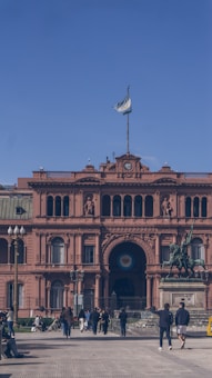 A vibrant pink historical building with a clock tower, adorned with multiple arches and sculptures. The Argentine flag is flying at the top. In the foreground, pedestrians are walking along the pathway, and a prominent equestrian statue stands to the side.