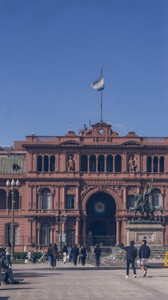 A vibrant pink historical building with a clock tower, adorned with multiple arches and sculptures. The Argentine flag is flying at the top. In the foreground, pedestrians are walking along the pathway, and a prominent equestrian statue stands to the side.