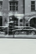 A blurred image of a car showroom, displaying a sleek sports car behind a large glass window. The reflection of a building with large windows and detailed brickwork can be seen on the glass.