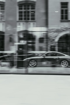 A blurred image of a car showroom, displaying a sleek sports car behind a large glass window. The reflection of a building with large windows and detailed brickwork can be seen on the glass.