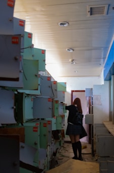 A young woman with red hair dressed in a school uniform explores a dimly lit corridor filled with rows of old, disheveled metal lockers. The scene appears to be in an abandoned or disused facility, suggested by the peeling paint and general disarray.