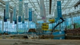 An abandoned indoor water park with decrepit structures, including a defunct yellow water slide and several hanging banners with text. The area is littered with debris and appears to be in disrepair. The large glass walls and ceiling let in natural light, revealing an overgrown and neglected landscape.