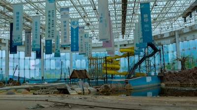 An abandoned indoor water park with decrepit structures, including a defunct yellow water slide and several hanging banners with text. The area is littered with debris and appears to be in disrepair. The large glass walls and ceiling let in natural light, revealing an overgrown and neglected landscape.