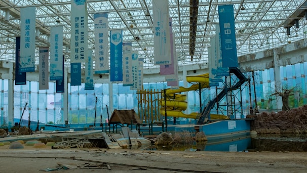 An abandoned indoor water park with decrepit structures, including a defunct yellow water slide and several hanging banners with text. The area is littered with debris and appears to be in disrepair. The large glass walls and ceiling let in natural light, revealing an overgrown and neglected landscape.