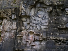 A detailed stone carving featuring intricate patterns and figures set against a rough, aged stone wall. The carvings include geometric shapes and what appears to be a face or mask. The stone has weathered textures, giving it an ancient and historical feel.