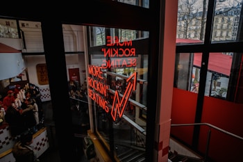 A neon sign reading 'MORE ROCKIN' SEATING' is prominently displayed on a glass window. Inside, there is a busy restaurant with several people, including staff behind a counter with red and white decor. Outside, trees without leaves and buildings are visible through large glass windows.