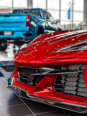 A close-up view of a red sports car front in a showroom with a blue pickup truck in the background. The focus is on the sleek, aerodynamic design and glossy finish of the sports car, emphasizing its modern and high-performance features. The blurred truck in the background contrasts with the sharp details of the car's bodywork.