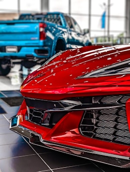 A close-up view of a red sports car front in a showroom with a blue pickup truck in the background. The focus is on the sleek, aerodynamic design and glossy finish of the sports car, emphasizing its modern and high-performance features. The blurred truck in the background contrasts with the sharp details of the car's bodywork.