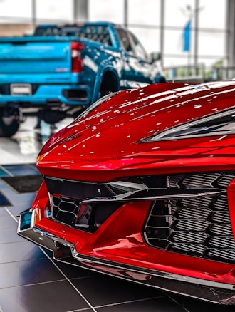 A close-up view of a red sports car front in a showroom with a blue pickup truck in the background. The focus is on the sleek, aerodynamic design and glossy finish of the sports car, emphasizing its modern and high-performance features. The blurred truck in the background contrasts with the sharp details of the car's bodywork.