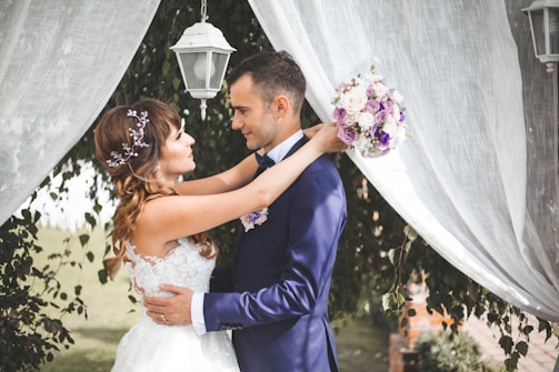 A couple stands close together, surrounded by white sheer curtains and foliage. The woman in a white lace dress holds a bouquet of purple and white flowers and gazes affectionately at the man dressed in a navy suit. An outdoor lantern hangs above them, adding to the romantic atmosphere.