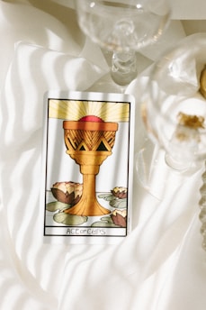 A tarot card featuring the Ace of Cups is placed on a softly lit, white fabric surface. The card illustrates a golden chalice with a red sphere above it, against a background of lily pads and blooming lotus flowers. Nearby, delicate glassware adds to the serene and reflective atmosphere.
