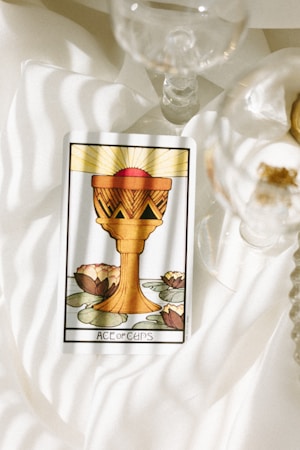 A tarot card featuring the Ace of Cups is placed on a softly lit, white fabric surface. The card illustrates a golden chalice with a red sphere above it, against a background of lily pads and blooming lotus flowers. Nearby, delicate glassware adds to the serene and reflective atmosphere.