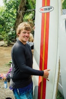 A person, smiling, holds a surfboard vertically. The surfboard has a red and orange stripe design with a logo, and the person is wearing a black rash guard and blue shorts. In the background, there are trees and some scattered belongings.