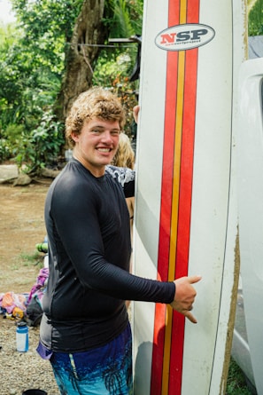 A person, smiling, holds a surfboard vertically. The surfboard has a red and orange stripe design with a logo, and the person is wearing a black rash guard and blue shorts. In the background, there are trees and some scattered belongings.