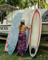 A woman in a swimsuit and patterned wrap stands beside three surfboards, one of which she is holding. There's a boat behind them with tropical palm trees in the background, creating a beach-like setting.