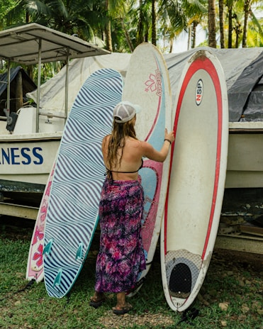 A woman in a swimsuit and patterned wrap stands beside three surfboards, one of which she is holding. There's a boat behind them with tropical palm trees in the background, creating a beach-like setting.