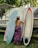 A woman in a swimsuit and patterned wrap stands beside three surfboards, one of which she is holding. There's a boat behind them with tropical palm trees in the background, creating a beach-like setting.