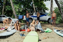 A group of seven people relax on a sandy beach surrounded by tropical trees. Surfboards are scattered on the ground, and a silver pickup truck is parked in the background with a hammock set up nearby. Some individuals are sitting on the sand enjoying food, while others are standing or engaged in conversation. Backpacks and beach gear are visible, adding to the laid-back and casual vibe.