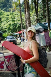 A person is smiling and holding a red surfboard under one arm while wearing a cap. They have a backpack slung over one shoulder and stand amidst trees and foliage in the background, along with other people and colorful tents.