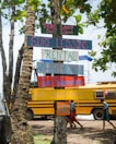 Colorful wooden signs are nailed to a tree, advertising services related to surfing, including a surf school, lessons, rentals, and repairs. In the background, there is a yellow school bus and a few people walking, with lush green trees overhead.