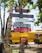 Colorful wooden signs are nailed to a tree, advertising services related to surfing, including a surf school, lessons, rentals, and repairs. In the background, there is a yellow school bus and a few people walking, with lush green trees overhead.
