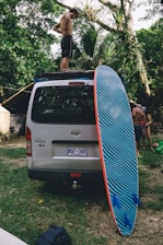 A person stands on the roof of a parked Toyota Hiace van in a lush, green environment. A large surfboard with blue and black stripes is leaning against the back of the van. Another person is visible in the background, crouching beside the van.