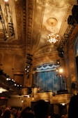 A grand interior of a theater or auditorium featuring an ornate ceiling with intricate patterns and a chandelier. Stage lights and rigging are visible. Curtains onstage are closed while audience members and staff are visible in the foreground.