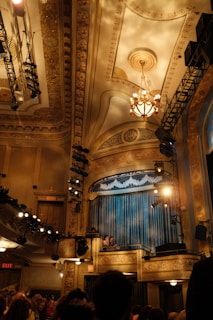A grand interior of a theater or auditorium featuring an ornate ceiling with intricate patterns and a chandelier. Stage lights and rigging are visible. Curtains onstage are closed while audience members and staff are visible in the foreground.