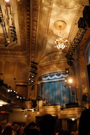 A grand interior of a theater or auditorium featuring an ornate ceiling with intricate patterns and a chandelier. Stage lights and rigging are visible. Curtains onstage are closed while audience members and staff are visible in the foreground.