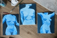 Three paintings of the human torso in shades of blue are laid out on a wooden surface. Each artwork focuses on different poses and angles with dynamic use of light and shadow. Paint brushes, a crumpled tissue, and a paint can are placed nearby, suggesting an artistic workspace.