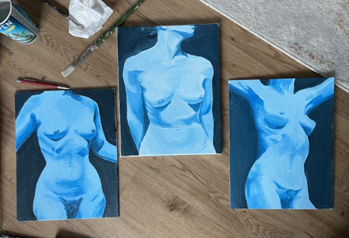Three paintings of the human torso in shades of blue are laid out on a wooden surface. Each artwork focuses on different poses and angles with dynamic use of light and shadow. Paint brushes, a crumpled tissue, and a paint can are placed nearby, suggesting an artistic workspace.