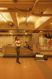 A person is singing in a subway station with a microphone, standing on a tiled floor. The surrounding area includes metal railings and an escalator. There are overhead lights that provide warm lighting. A sign indicates the location as 34 St & 6 Avenue.