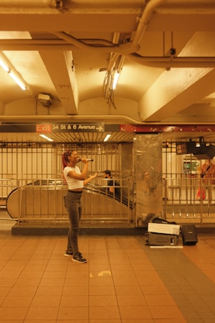 A person is singing in a subway station with a microphone, standing on a tiled floor. The surrounding area includes metal railings and an escalator. There are overhead lights that provide warm lighting. A sign indicates the location as 34 St & 6 Avenue.