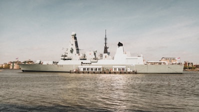A large, modern military ship is docked by the waterfront, displaying advanced radar and communication equipment on its deck. Several flags are visible atop the vessel, while the surroundings include urban buildings and a mostly clear sky.