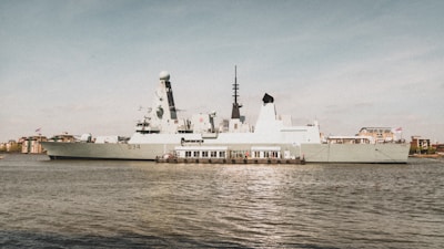 A large, modern military ship is docked by the waterfront, displaying advanced radar and communication equipment on its deck. Several flags are visible atop the vessel, while the surroundings include urban buildings and a mostly clear sky.
