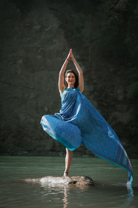 A person is gracefully balancing in a yoga pose on a rock surrounded by water. They are wearing a flowing blue garment and have their hands raised above their head in a prayer position. The background is a dark, natural setting.