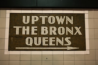 A mosaic sign on a tiled wall features the words 'UPTOWN THE BRONX QUEENS' with an arrow pointing to the right. The letters are made up of white tiles set against a darker tiled background, creating a clear contrast.
