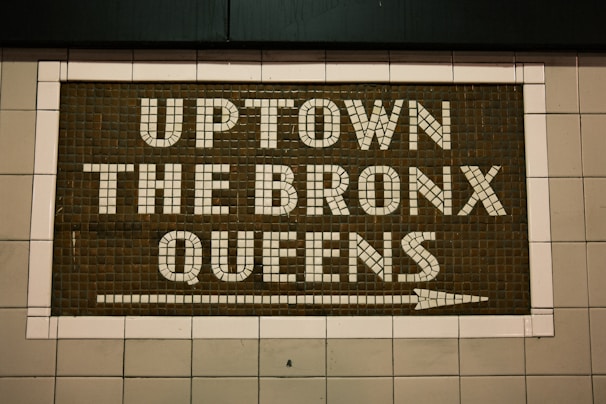 A mosaic sign on a tiled wall features the words 'UPTOWN THE BRONX QUEENS' with an arrow pointing to the right. The letters are made up of white tiles set against a darker tiled background, creating a clear contrast.