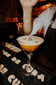 A cocktail in a martini glass garnished with coffee beans is placed on a bar mat. The arm of a bartender in a white shirt and a dark apron is visible, gesturing above the drink. The background is dimly lit, providing a warm and cozy atmosphere typical of a bar setting.