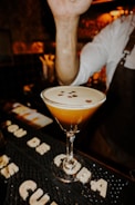 A cocktail in a martini glass garnished with coffee beans is placed on a bar mat. The arm of a bartender in a white shirt and a dark apron is visible, gesturing above the drink. The background is dimly lit, providing a warm and cozy atmosphere typical of a bar setting.