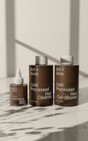 Three brown bottles with white caps are arranged on a light surface. The bottles are labeled as 'Act + Acre' and include 'Cold Processed Scalp Detox,' 'Cold Processed Hair Cleanse,' and 'Cold Processed Hair Conditioner.' Shadows of nearby objects are visible on the surface.