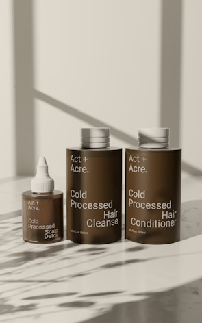 Three brown bottles with white caps are arranged on a light surface. The bottles are labeled as 'Act + Acre' and include 'Cold Processed Scalp Detox,' 'Cold Processed Hair Cleanse,' and 'Cold Processed Hair Conditioner.' Shadows of nearby objects are visible on the surface.