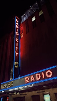 An illuminated marquee and vertical sign of Radio City Music Hall, featuring bright neon lights in red and blue, advertising a 'Christmas Spectacular' against a dark city backdrop.