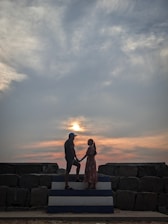 Two people stand facing each other on a stepped platform, silhouetted against a dramatic sky with clouds and a setting sun. They appear to be holding hands, creating a romantic and intimate atmosphere.