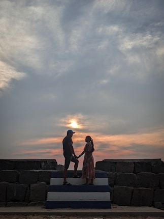 Two people stand facing each other on a stepped platform, silhouetted against a dramatic sky with clouds and a setting sun. They appear to be holding hands, creating a romantic and intimate atmosphere.