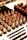 An assortment of lipsticks in various shades of red and brown is arranged in an organized manner. The lipsticks have black bases and gold holders, with some in open position revealing the color. Below them are makeup pencils with black and golden details, laid out neatly on a white surface.