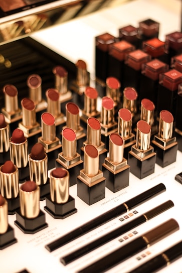 An assortment of lipsticks in various shades of red and brown is arranged in an organized manner. The lipsticks have black bases and gold holders, with some in open position revealing the color. Below them are makeup pencils with black and golden details, laid out neatly on a white surface.