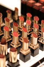 A display of various lipsticks, each housed in a gold and black casing, arranged neatly in rows. The lipsticks come in different shades of red and brown, and the background is slightly blurred, emphasizing the lipsticks.