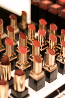 A display of various lipsticks, each housed in a gold and black casing, arranged neatly in rows. The lipsticks come in different shades of red and brown, and the background is slightly blurred, emphasizing the lipsticks.